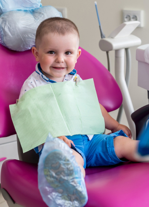 A child sits smiling in a dentist's chair with dental equipment around. The child wears a green bib and blue shorts.