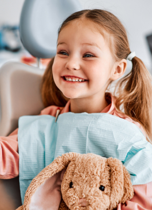 A smiling child with pigtails sits in a chair, holding a stuffed bunny, wearing a bib, seemingly at a dental office.
