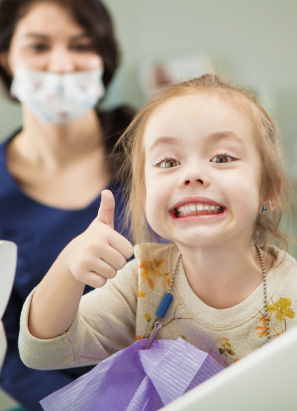 A smiling child gives a thumbs-up while a person in a mask stands in the background, likely in a dental clinic or medical setting.