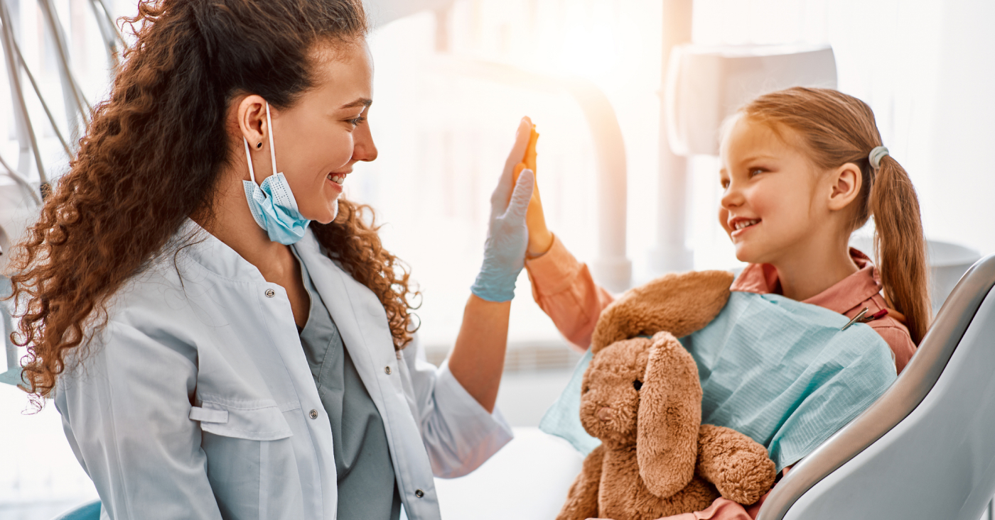 A person in a dental office shares a high-five with a young girl holding a stuffed bunny, both smiling brightly.