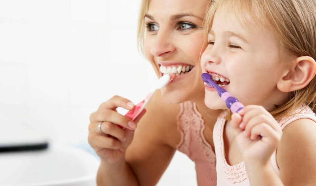 A person and a child are smiling while brushing their teeth together, wearing matching pink tops in a bright bathroom setting.