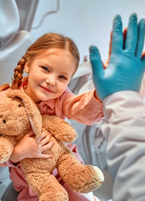 A child holding a teddy bear gives a high-five to a gloved person in a medical or dental setting, smiling brightly.