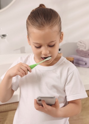 A child in a bathroom brushes their teeth with a green toothbrush while looking at a smartphone. Towels are stacked in the background.