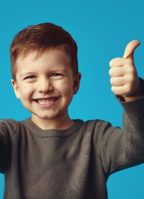 A child smiling broadly, giving a thumbs-up gesture, wearing a grey sweater against a plain blue background. No landmarks or historical buildings are visible.