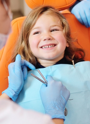 A child with long hair smiles in a dental chair while a person wearing gloves appears to be preparing for a dental procedure.