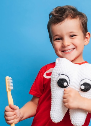 A child wearing a red shirt holds a toothbrush and a plush tooth-shaped toy, smiling against a blue background. No adults, landmarks, or historical buildings present.
