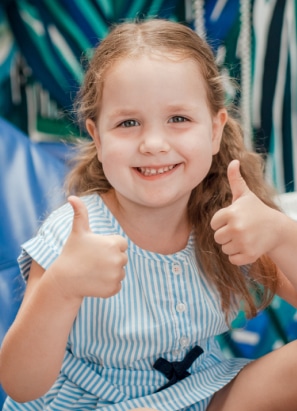 A smiling child with brown hair gives two thumbs up, wearing a blue and white striped dress. A patterned background is behind her.