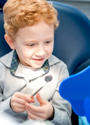 A child with curly hair happily holds a dental mirror while sitting on a blue dental chair in a brightly lit room.