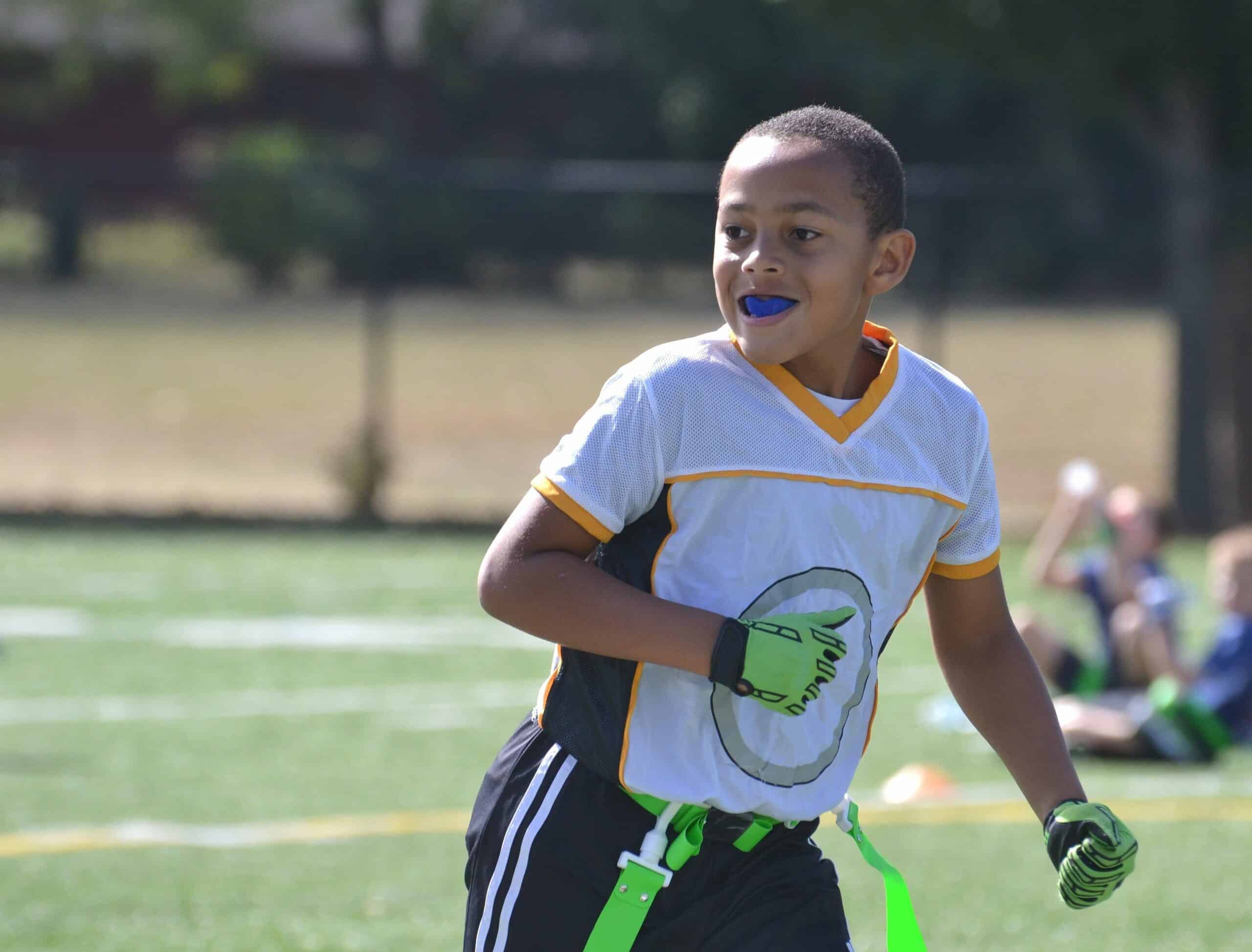 A child plays flag football on a sunny day, wearing a white jersey and gloves. Others are in the background on the field.