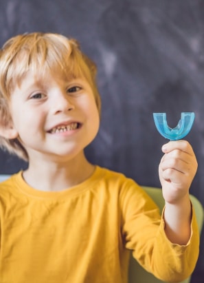 A smiling child wearing a yellow shirt holds a blue mouthguard, posing against a dark, blurred background. No landmarks or historical buildings visible.