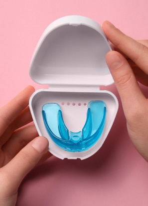A person holds an open white case containing a blue dental mouthguard against a pink background. Hands are positioned on either side of the case.