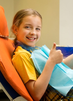 A child in a dentist's chair gives a thumbs up while wearing a yellow shirt and a blue bib, smiling brightly at the camera.