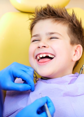 A child is laughing while a person wearing blue gloves examines their teeth in a dental chair. No recognizable landmarks or historical buildings are visible.
