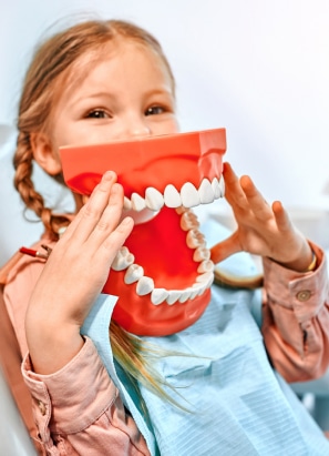 A child holds a large model of teeth, smiling playfully. She is wearing a dental bib, suggesting a dentist's office setting.
