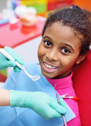 A child smiles while waiting for a dental check-up. A person, wearing gloves, holds dental instruments near the child's mouth. Bright colors in background.