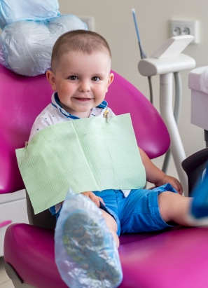 A child sits on a pink dental chair, wearing a dental bib, smiling, at a dental office, surrounded by professional equipment and supplies.