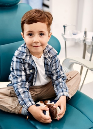 A child with brown hair sits cross-legged on a teal dental chair, smiling, in a brightly lit dental office with equipment in the background.