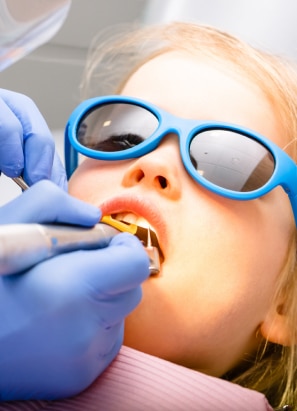 A child wearing blue sunglasses receives dental care from a gloved person. Reflective tools and a dental chair are visible in the background.