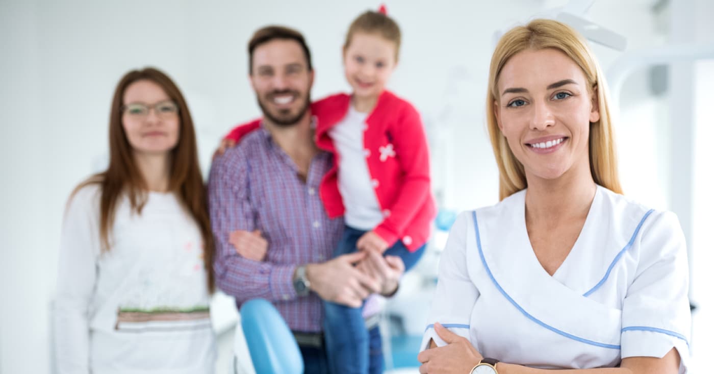 A person in a nurse uniform smiles while three people, including a child, stand in the background of a bright, modern room.