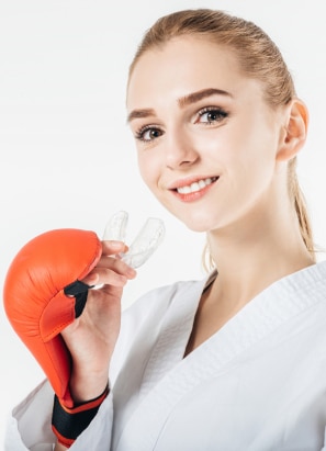 A person in martial arts attire holds a red boxing glove and a mouthguard, smiling softly. The background is plain white.