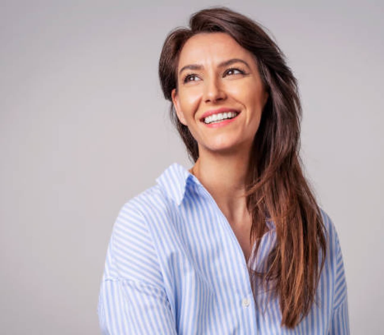 A person with long hair and a striped shirt smiles and looks away from the camera against a plain background.