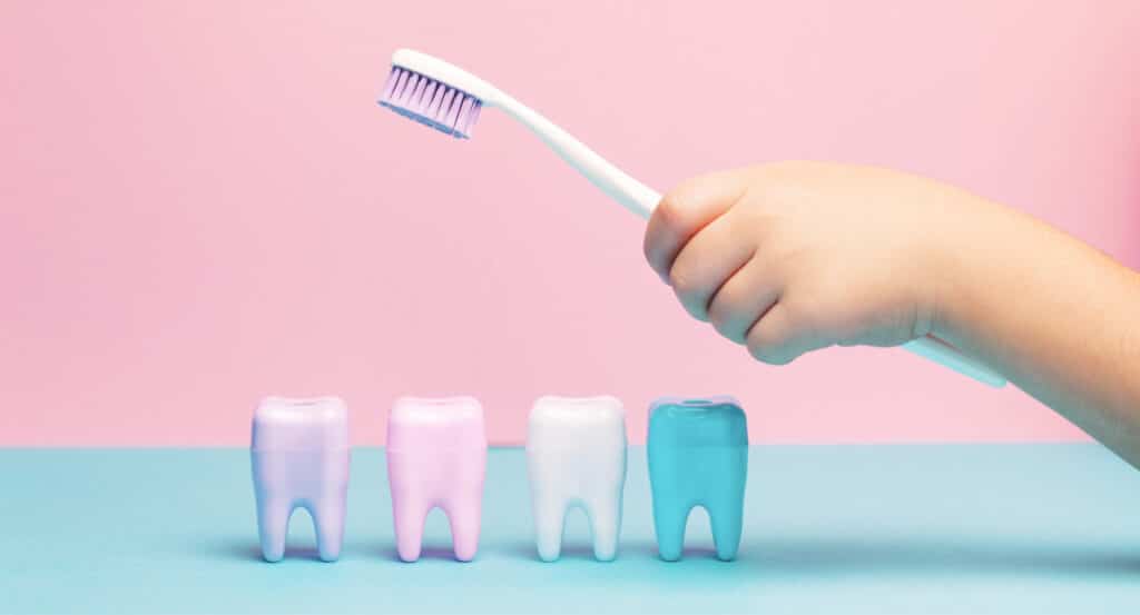 A hand holding a toothbrush is poised above rows of colorful tooth figurines against a pastel pink and blue background.