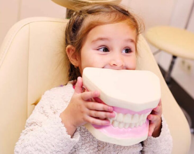 Young girl in dentist chair holding up a giant, plastic replica of teeth.
