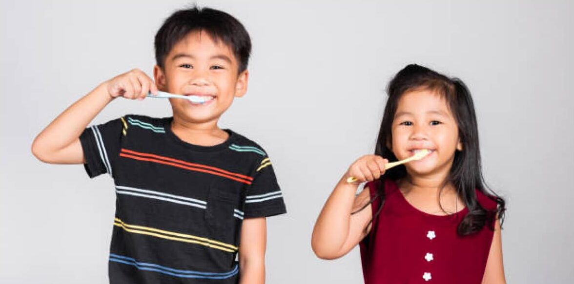 Two young children with brown hair, brushing their teeth in front of a plain gray background.