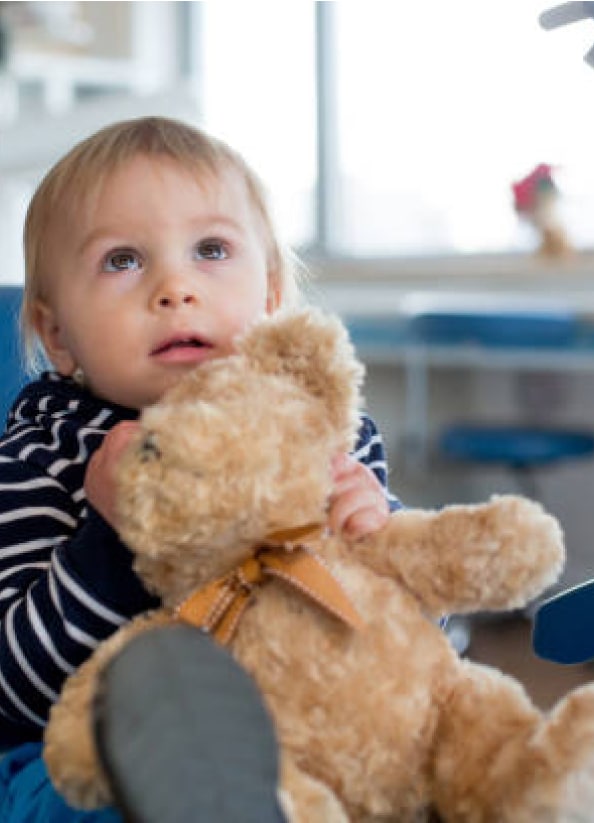 A child with light hair holds a brown teddy bear, gazing upward indoors. The background features blurred furniture and a window. No landmarks visible.