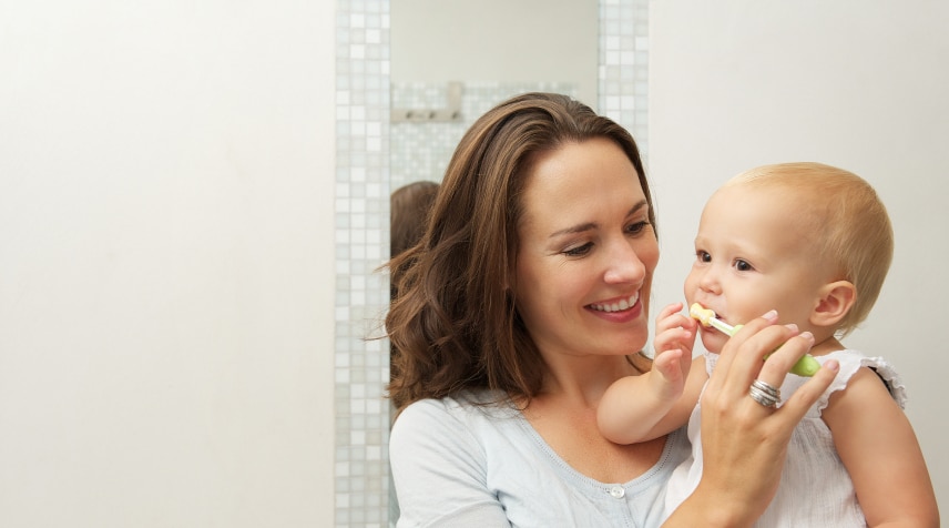 A person smiles while helping a baby with toothbrushing in a bright bathroom with tiled walls. The atmosphere is warm and nurturing.