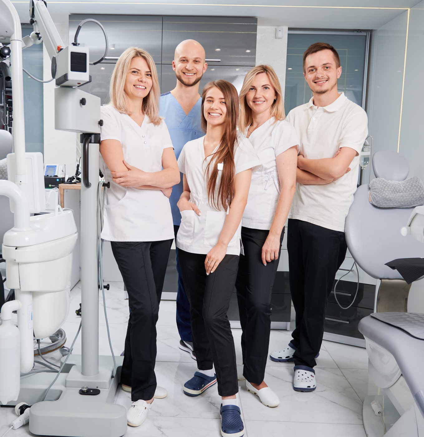Five persons in a dental clinic, standing and smiling, wearing professional attire. Dental equipment and chairs are visible in the background.