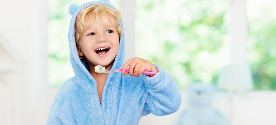 A child in a blue bear-themed robe is happily brushing their teeth with a pink toothbrush, set against a bright, blurred indoor background.