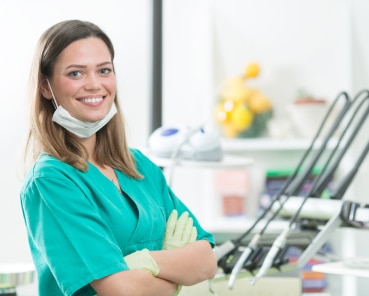 A person in scrubs and gloves, smiling in a medical or dental office setting with equipment and supplies in the background.