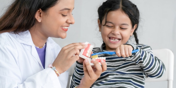 A person in a white coat assists a smiling child in brushing a dental model, demonstrating proper teeth brushing technique in a clinical setting.