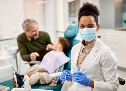 A person in a dental clinic smiles at the camera, while another person and a child are seated in the background during a dental check-up.