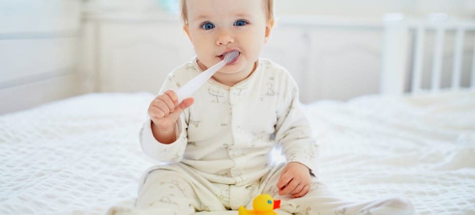 A baby in pajamas sits on a white bed, chewing on a toothbrush, with a yellow rubber duck toy nearby.