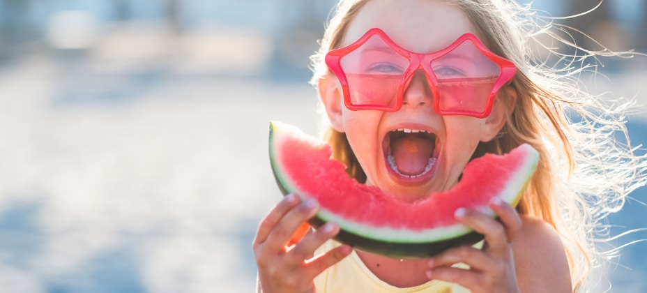 A child wearing red star-shaped sunglasses holds a slice of watermelon and laughs in a bright outdoor setting, possibly at the beach.