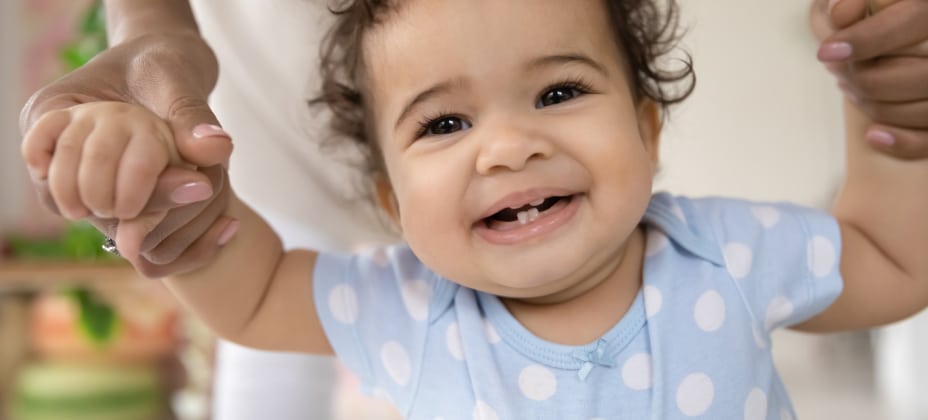 A smiling baby in a blue polka-dotted outfit, supported by a person's hands in a bright indoor setting. No landmarks or historical buildings visible.