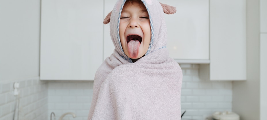 A child in a cozy gray towel with ears sticks out their tongue playfully in a bright kitchen with white cabinets and subway tiles.