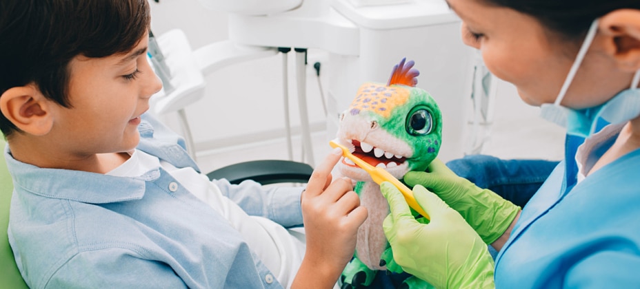 A child and a person, likely a dentist, are brushing a toy dinosaur's teeth together, possibly demonstrating dental care techniques in a dental office.
